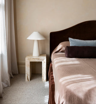 Bedroom with a burgundy velvet bed, stone table, and lamp against a beige wall.