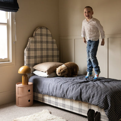 Child standing on a bed in a bedroom with plaid headboard and checkered bedding.