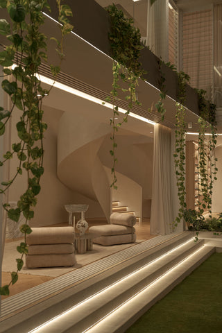 A modern architectural interior with a curved staircase, sculptural beige occasional chairs by create estate and hanging green vines, with warm ambient lighting along white steps.