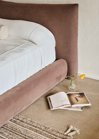 Brown upholstered bed head with a white bed and a vase with a single orange poppy flower on a chunky rug.