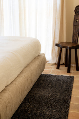 Beige ruched bed base next to a wooden chair on a dark rug in a room with light-colored walls and curtains.
