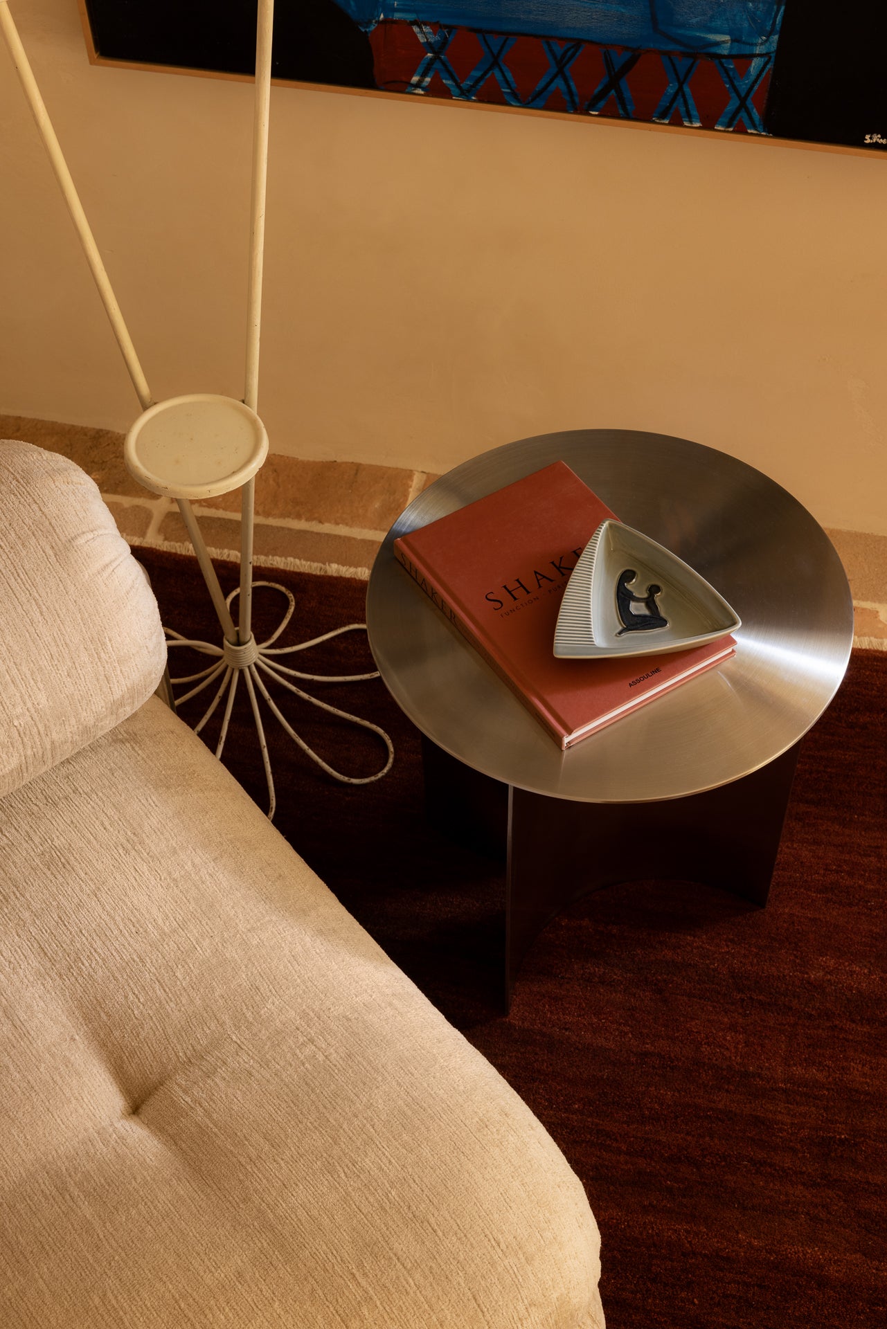 Round stainless steel side table with a red book and white object next to a beige chair.