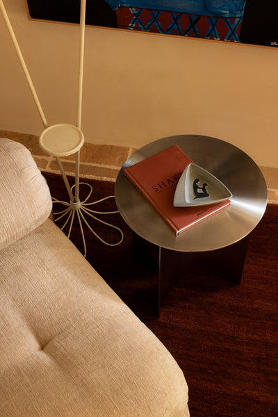 Round stainless steel side table with a red book and white object next to a beige chair.